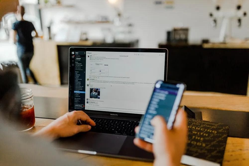 slack-app-laptop A person works on a laptop displaying a messaging app while holding a smartphone, with a cafe background.