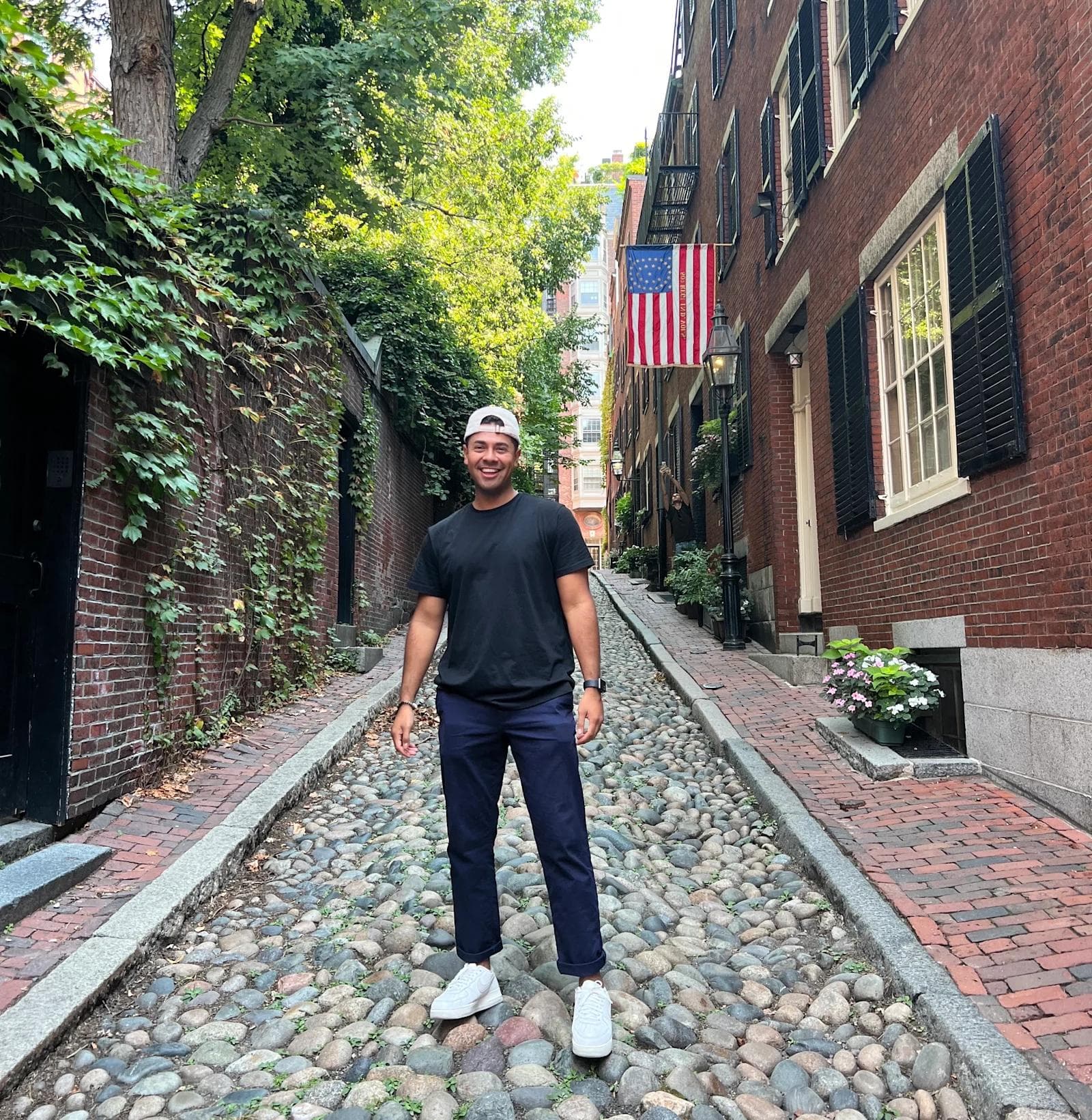 Eric BTB 6 Person in a black t-shirt and cap stands on a cobblestone street lined with brick buildings and greenery, an American flag visible.