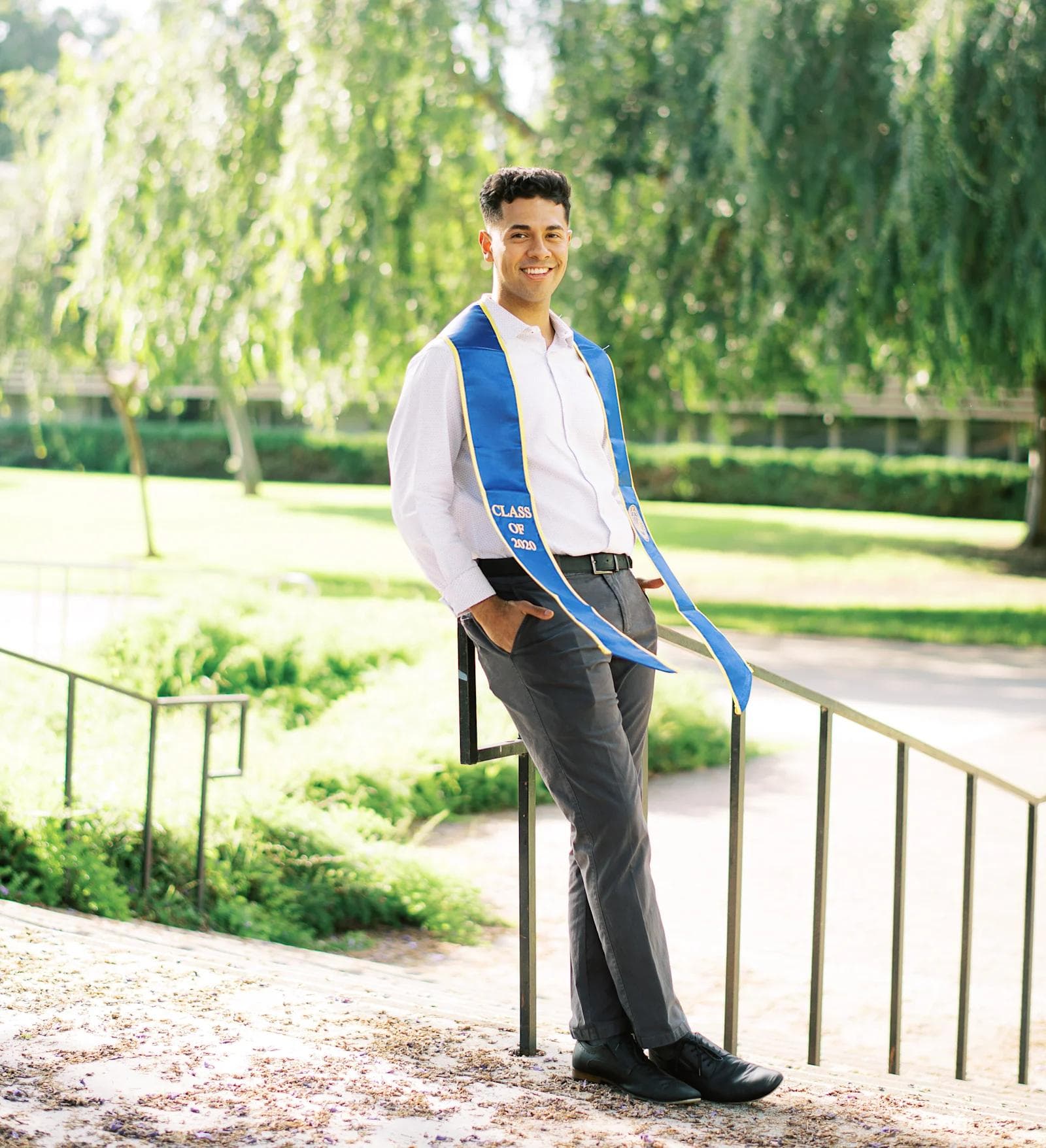 Eric BTB Picture 1 A smiling male graduate in a blue sash stands near steps in a green park, celebrating the Class of 2023.