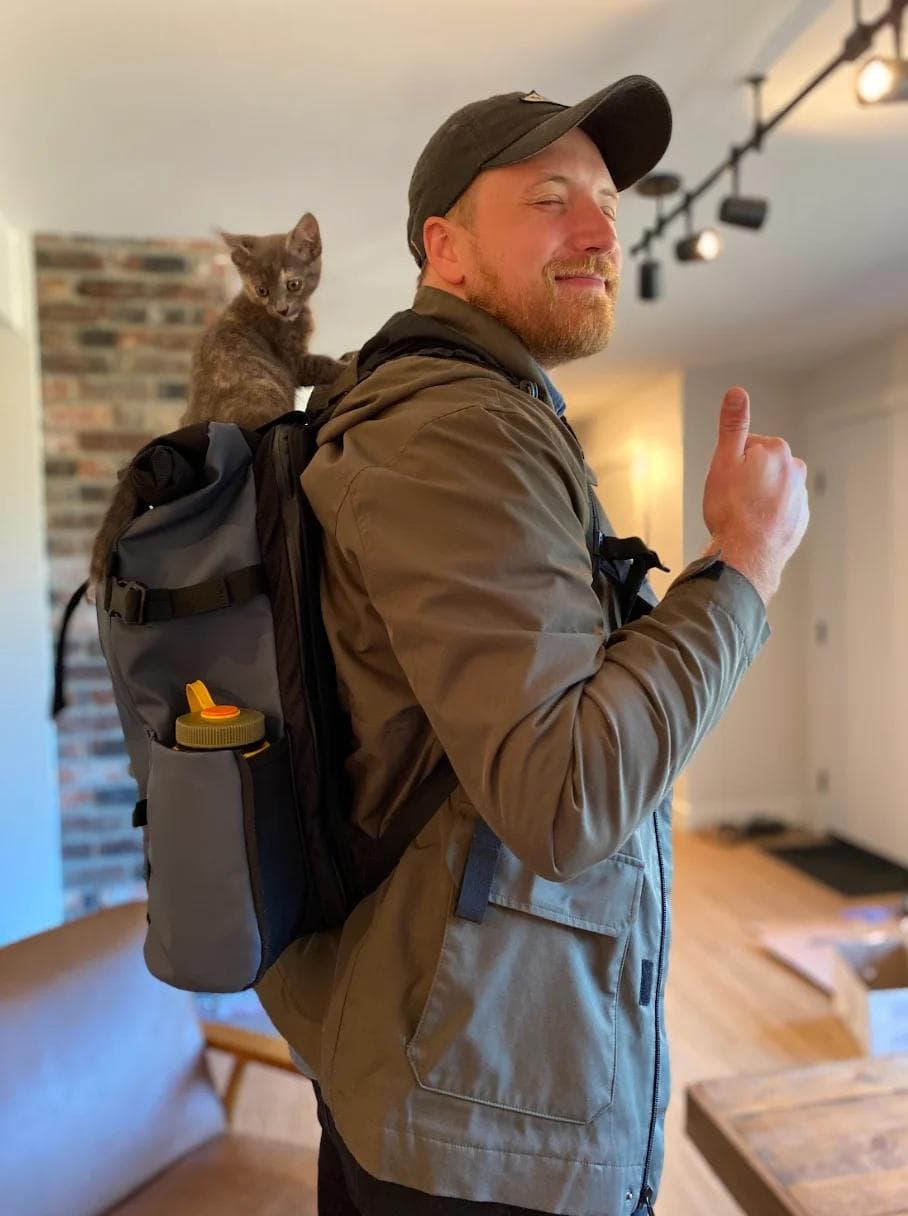 Joel BTB Picture 2 A man smiles and gives a thumbs up, with a gray kitten perched on his backpack.