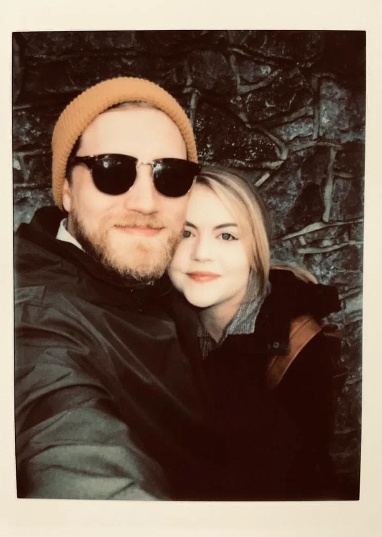 Joel BTB Picture 4 A couple poses together, smiling, with a stone wall in the background. The man wears sunglasses and a beanie.