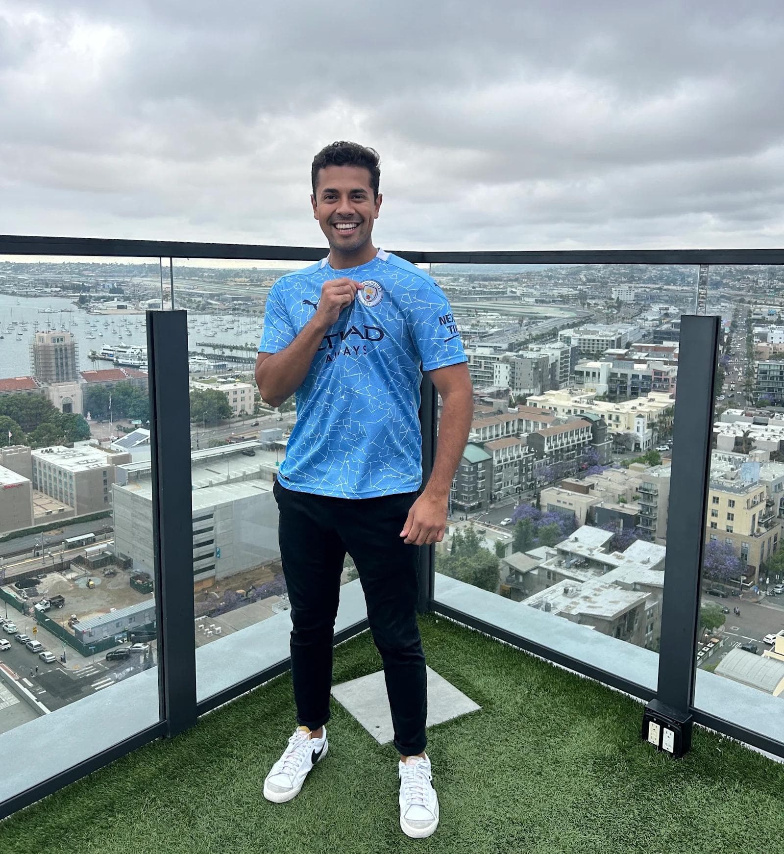 Eric BTB Picture 2 Man in a blue soccer jersey smiles while standing on a rooftop with a city skyline and cloudy sky in the background.