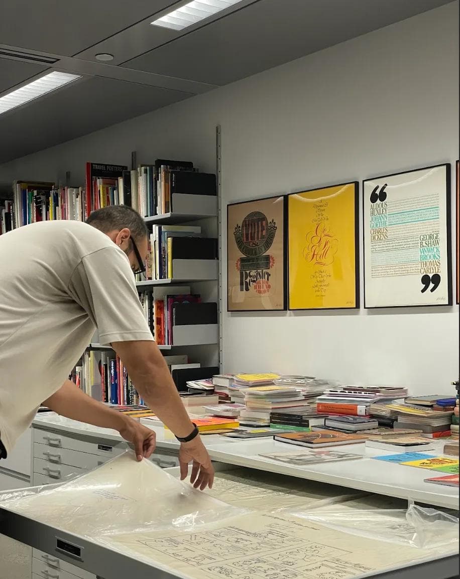 BTB Jordan Layao Image 2 A person examines large sheets of artwork on a table in a bright, modern workspace surrounded by books and posters.